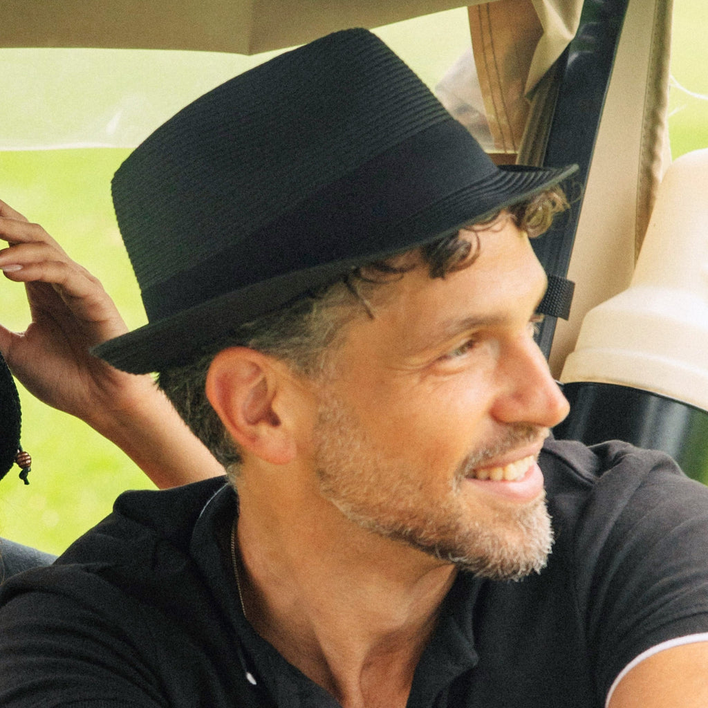 A man wearing a black fedora hat and a black polo shirt, smiling while seated in a vehicle.