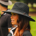 A woman in profile wearing a black woven wide-brim hat with small decorative beads on the band, photographed outdoors with a blurred green background and another person partially visible behind her.