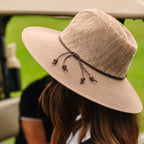 A beige woven sun hat with a dark brown band and drawstring, shown on a person in a golf cart with a blurred green background.