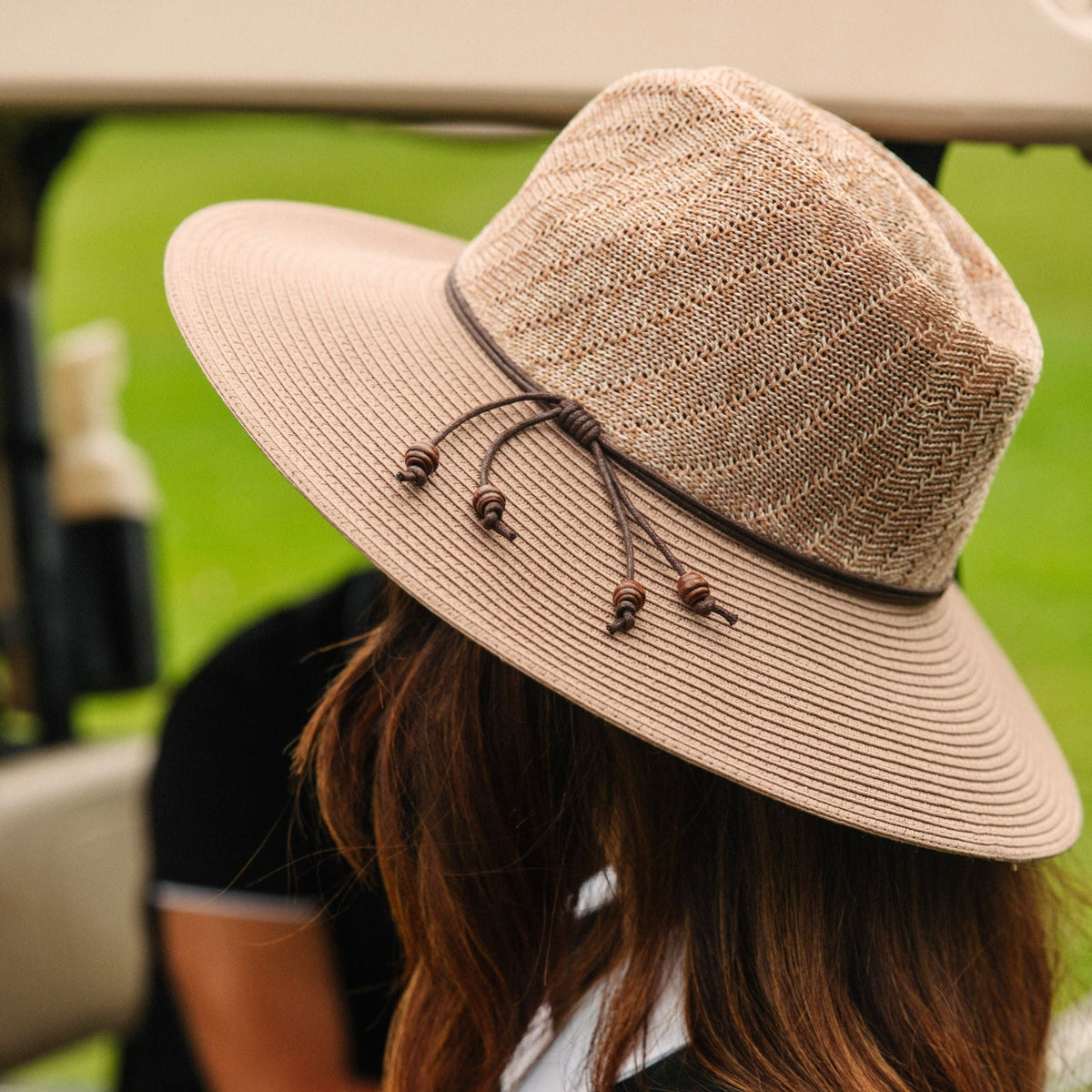A beige woven sun hat with a dark brown band and drawstring, shown on a person in a golf cart with a blurred green background.