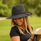 A woman wearing a black woven sun hat with a curved brim and a decorative cord detail, photographed outdoors in a grassy park setting.