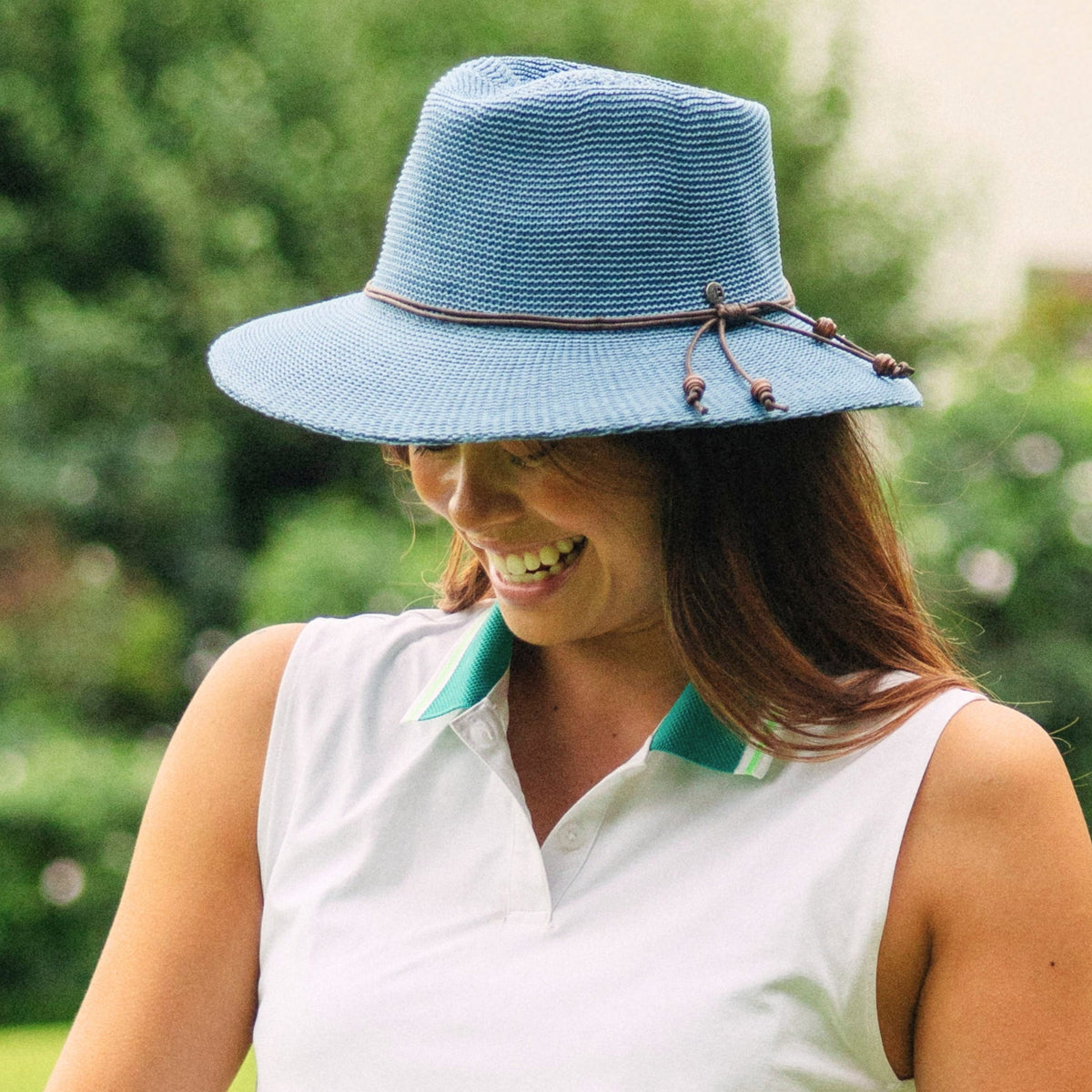 A woman outdoors wearing a light blue woven sun hat with a brown rope detail and a white sleeveless polo shirt with green collar accents.