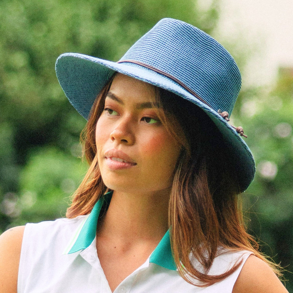 A woman wearing a light blue woven sun hat with a dark band and a white sleeveless top with a teal collar, photographed outdoors against a blurred green background.